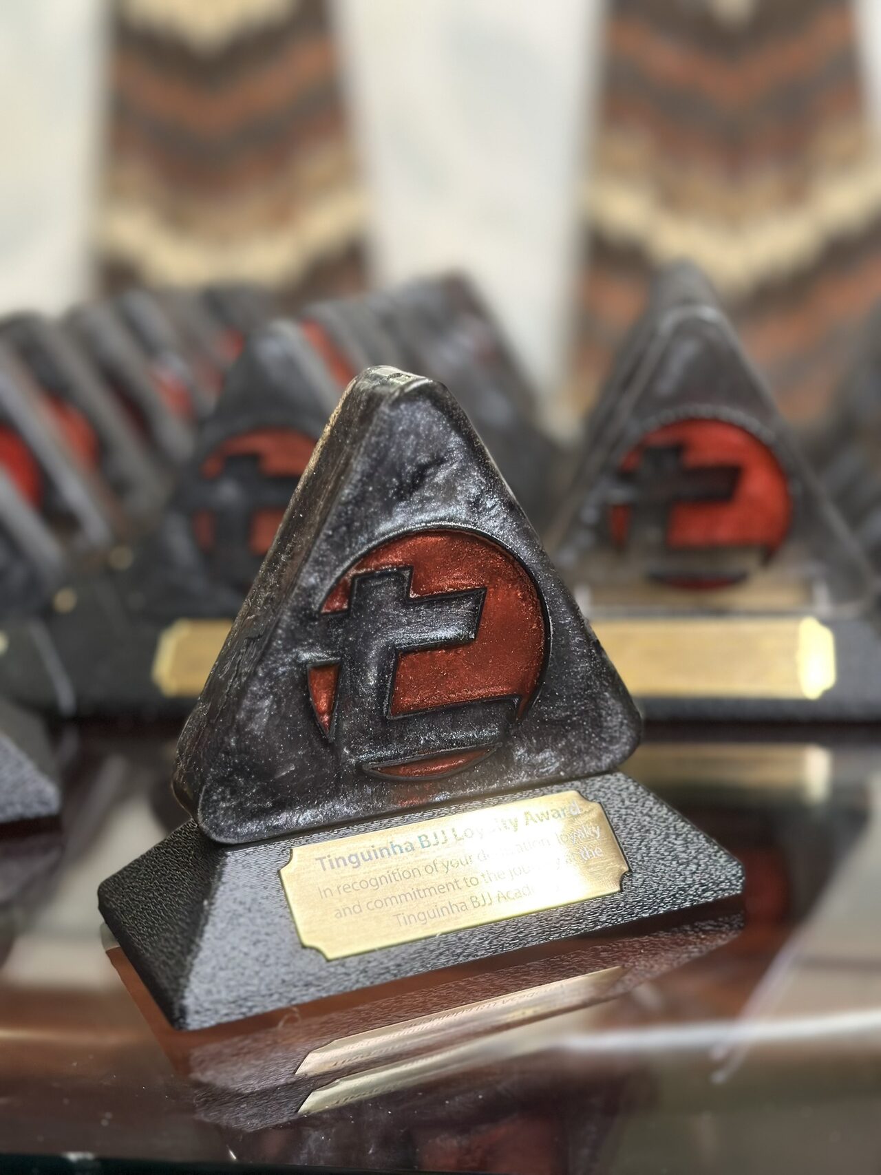 A triangular award with a red resin logo in a black marbled body, standing on a textured black base with a brass plate reading Tinguinha BJJ Loyalty Award. Rows of matching awards are blurred behind it on a glass table.