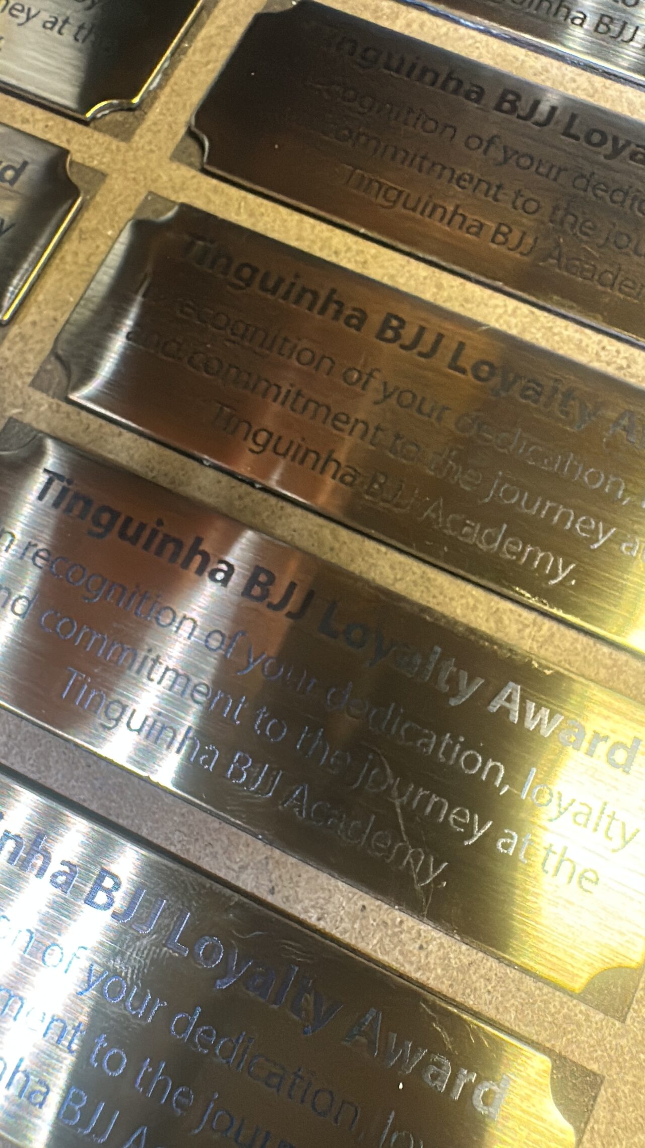 Close-up of engraved brass plates seated in a cut jig, each reading Tinguinha BJJ Loyalty Award with two lines of text below.